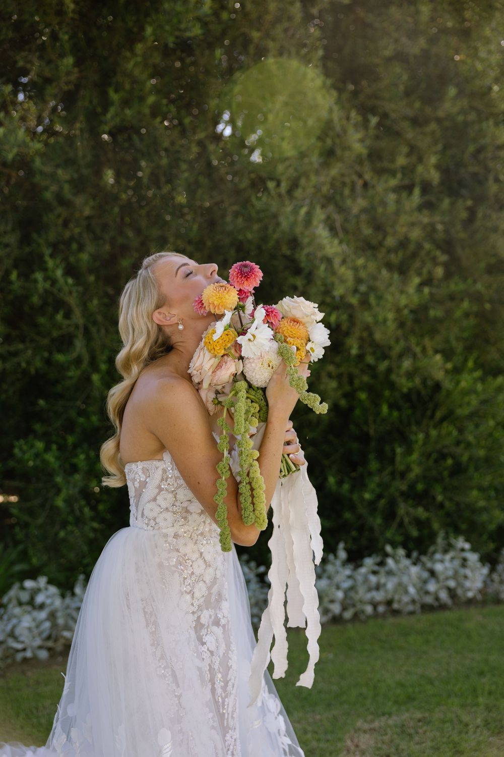 Bride in lace gown smelling colorful wedding bouquet with cascading ribbons outdoors in garden setting.