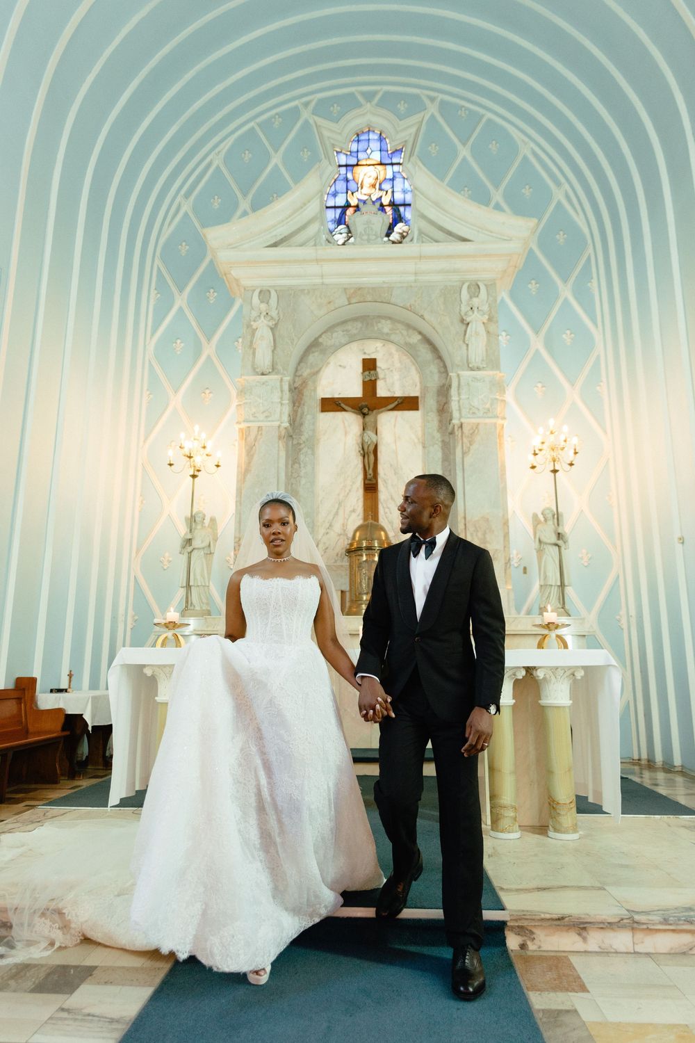 Bride and groom holding hands at church altar, bride in white gown, groom in black tuxedo, crucifix backdrop.