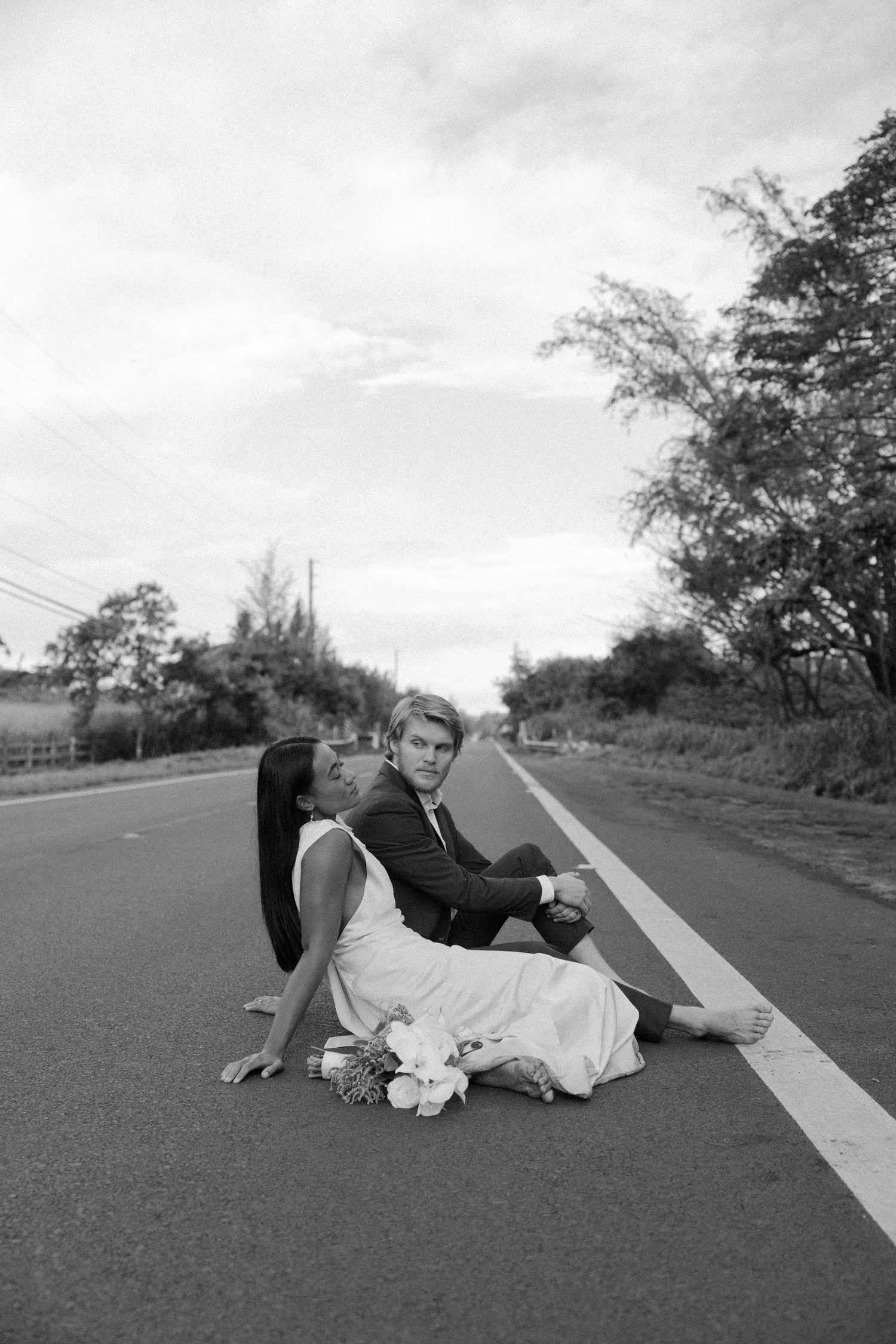 A couple in formal wear sitting on a road with mountains in the distance.