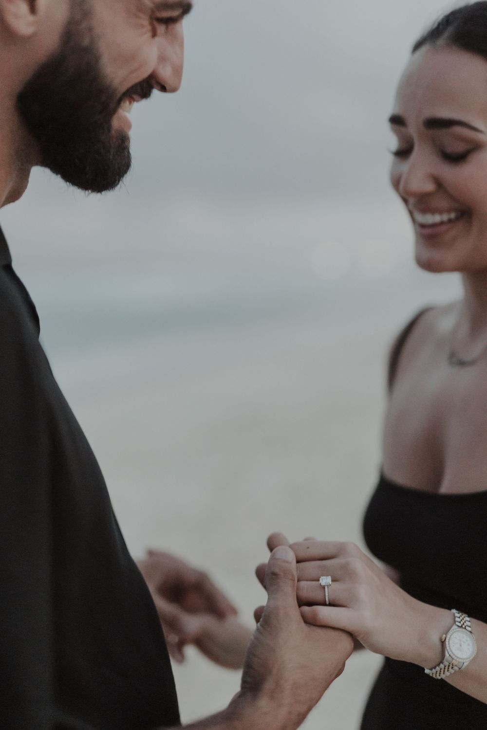 Smiling couple sharing a romantic engagement moment on the beach, with a diamond ring being placed on her finger.