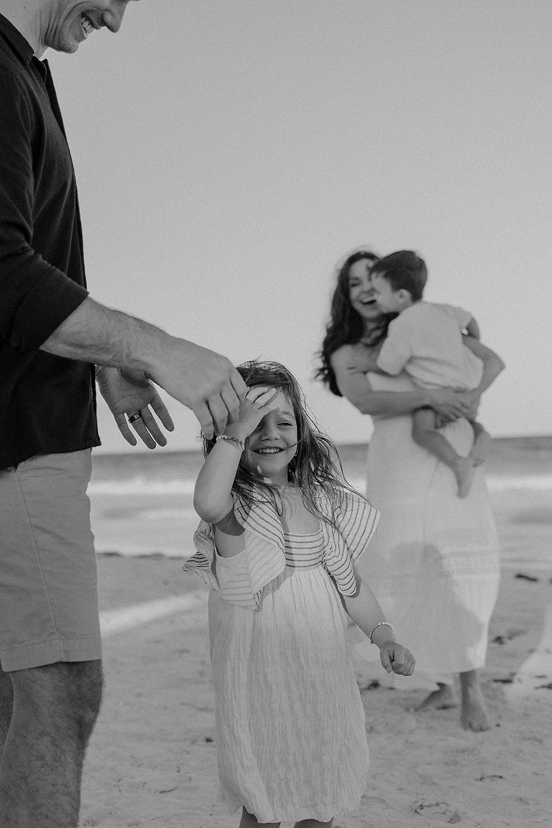 Black and white beach photo of a laughing girl in a white dress with her family on a sunny shore.