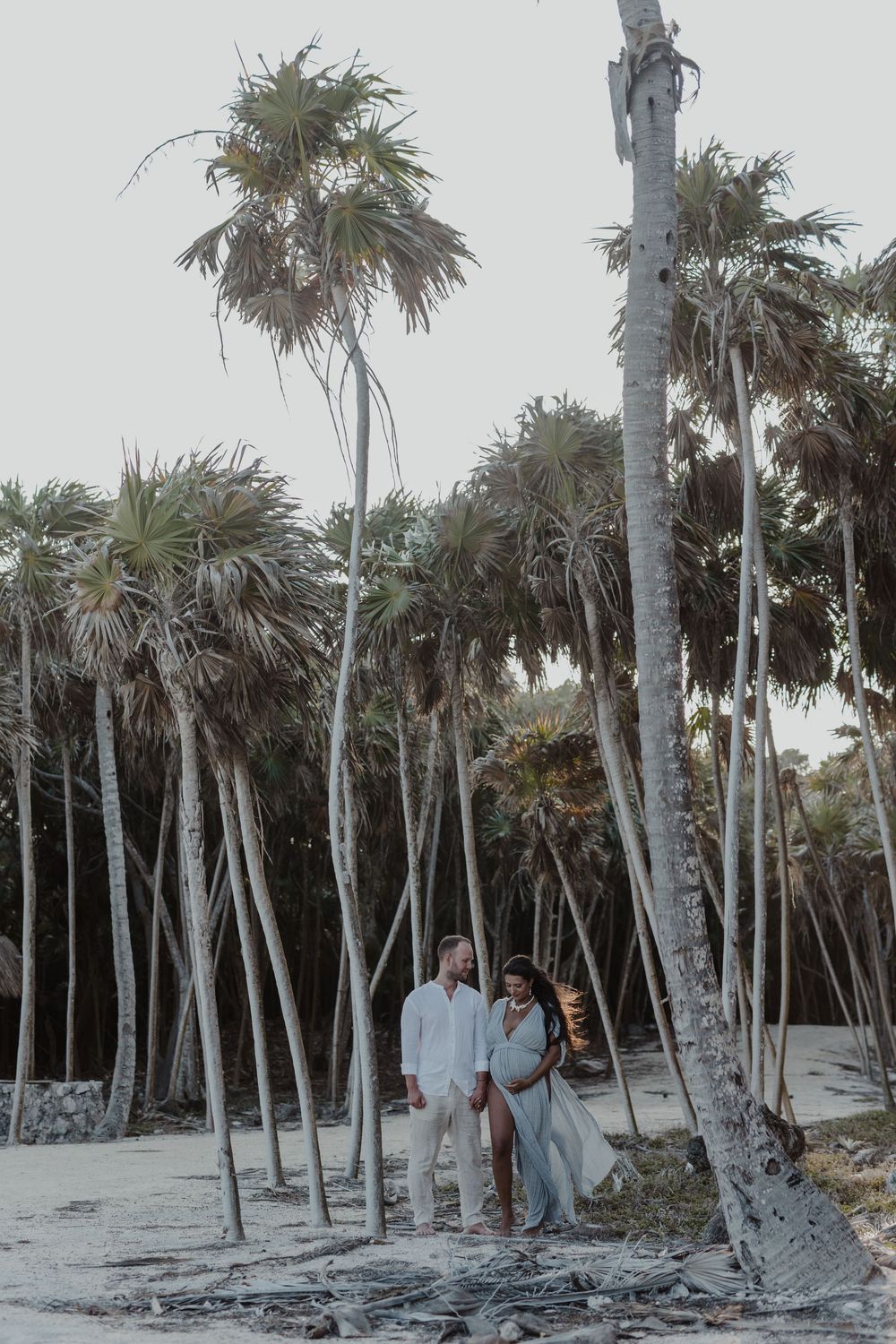 Couple standing among tall palm trees on a sandy beach, woman in flowing dress, man in white, tropical maternity photo.