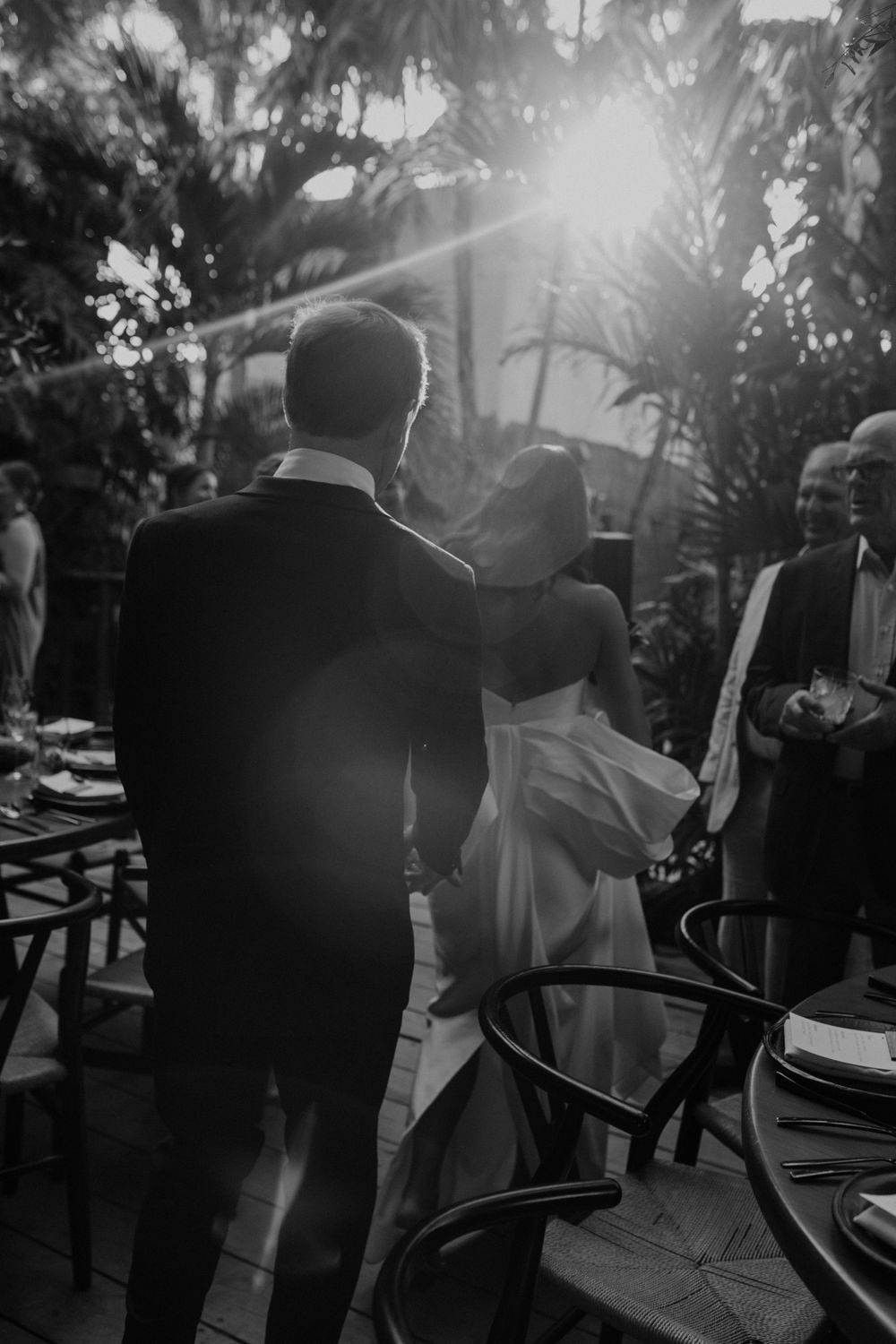Black and white photo of bride and groom walking hand in hand at outdoor wedding reception with sunlight streaming through tropical palms.