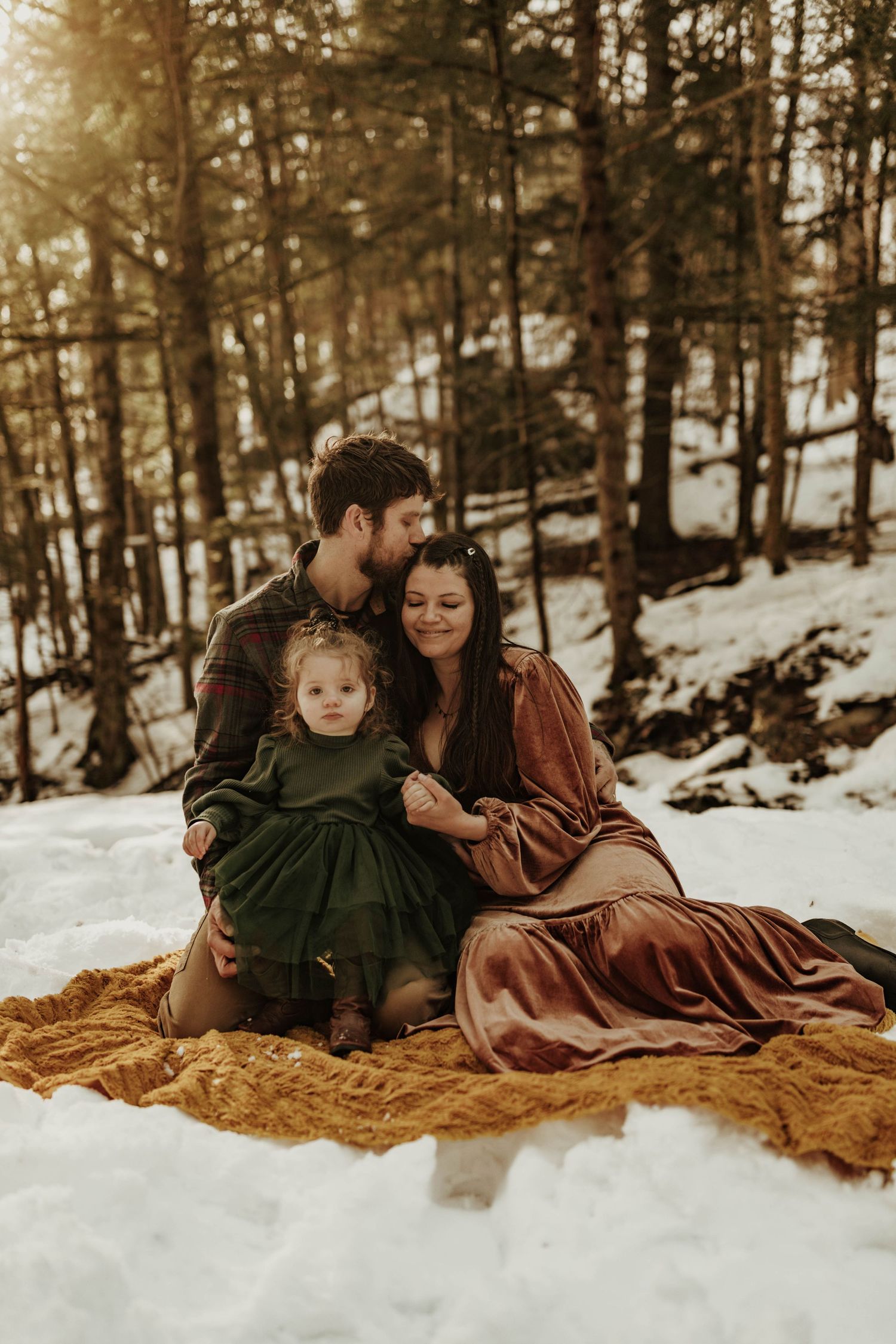 A family sits on a golden blanket in the snow during a winter forest photoshoot.