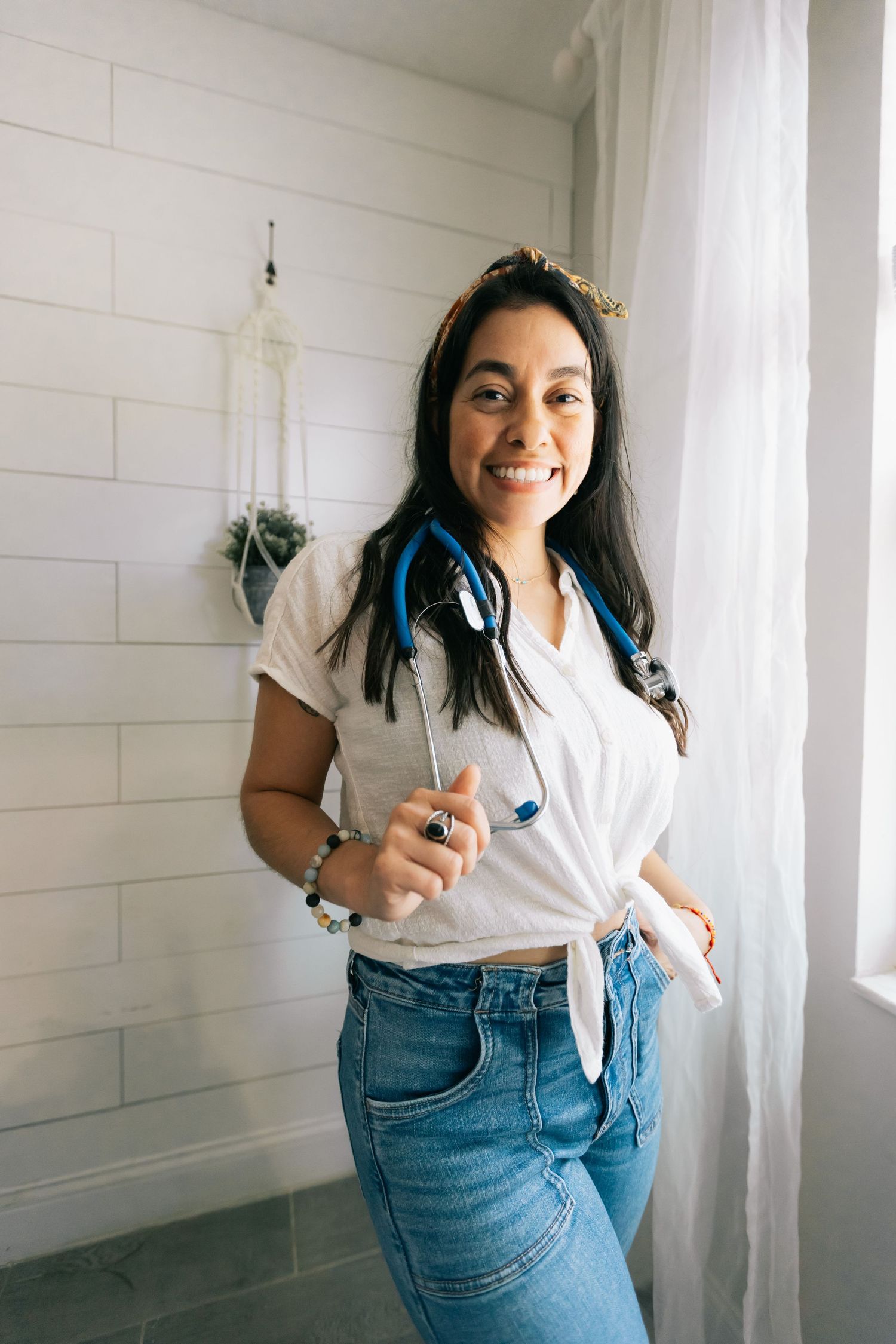 Smiling woman in a knotted white shirt and jeans with a blue stethoscope around her neck stands in a bright room.