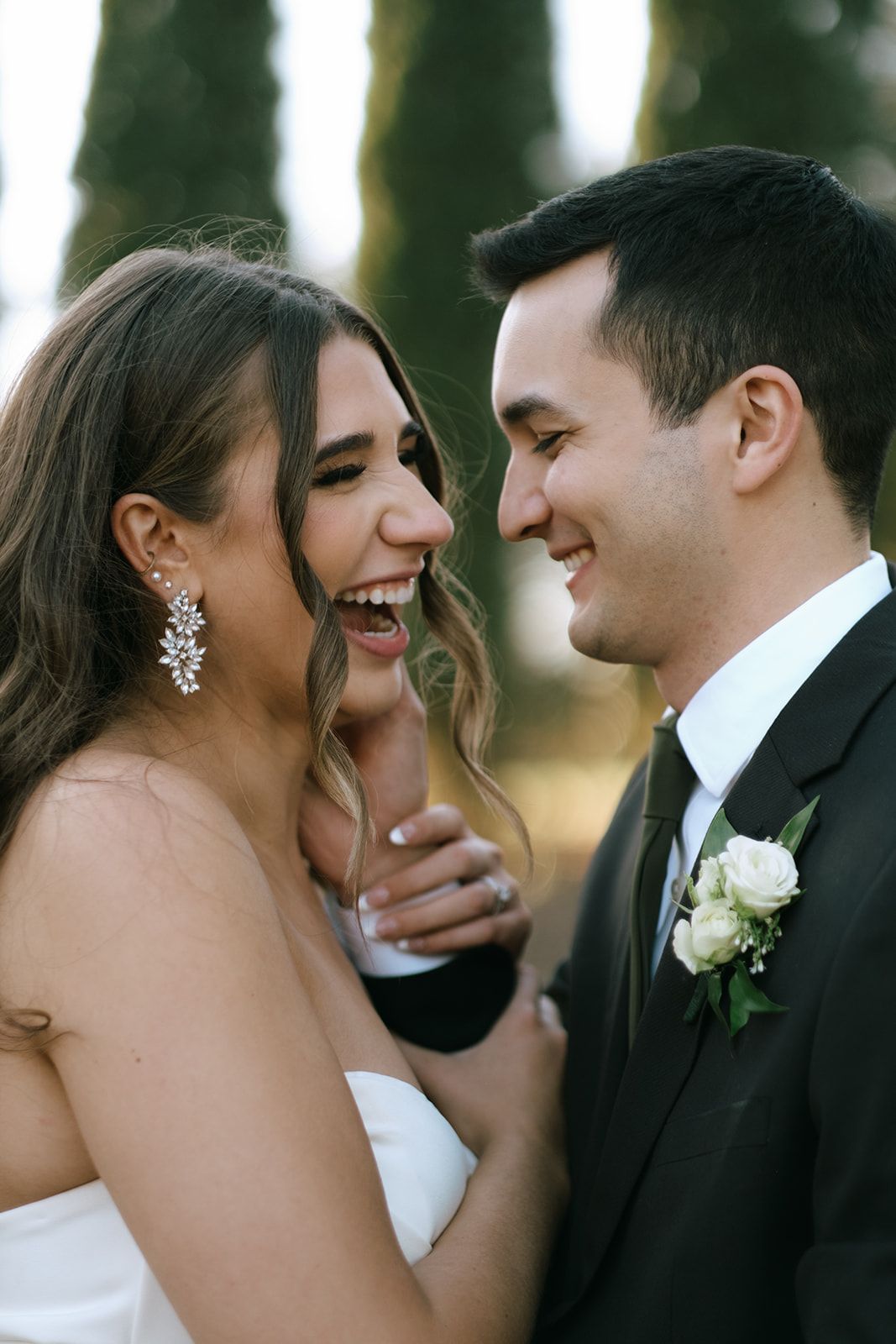 Laughing bride with crystal earrings and groom in black tuxedo share joyful moment outdoors.