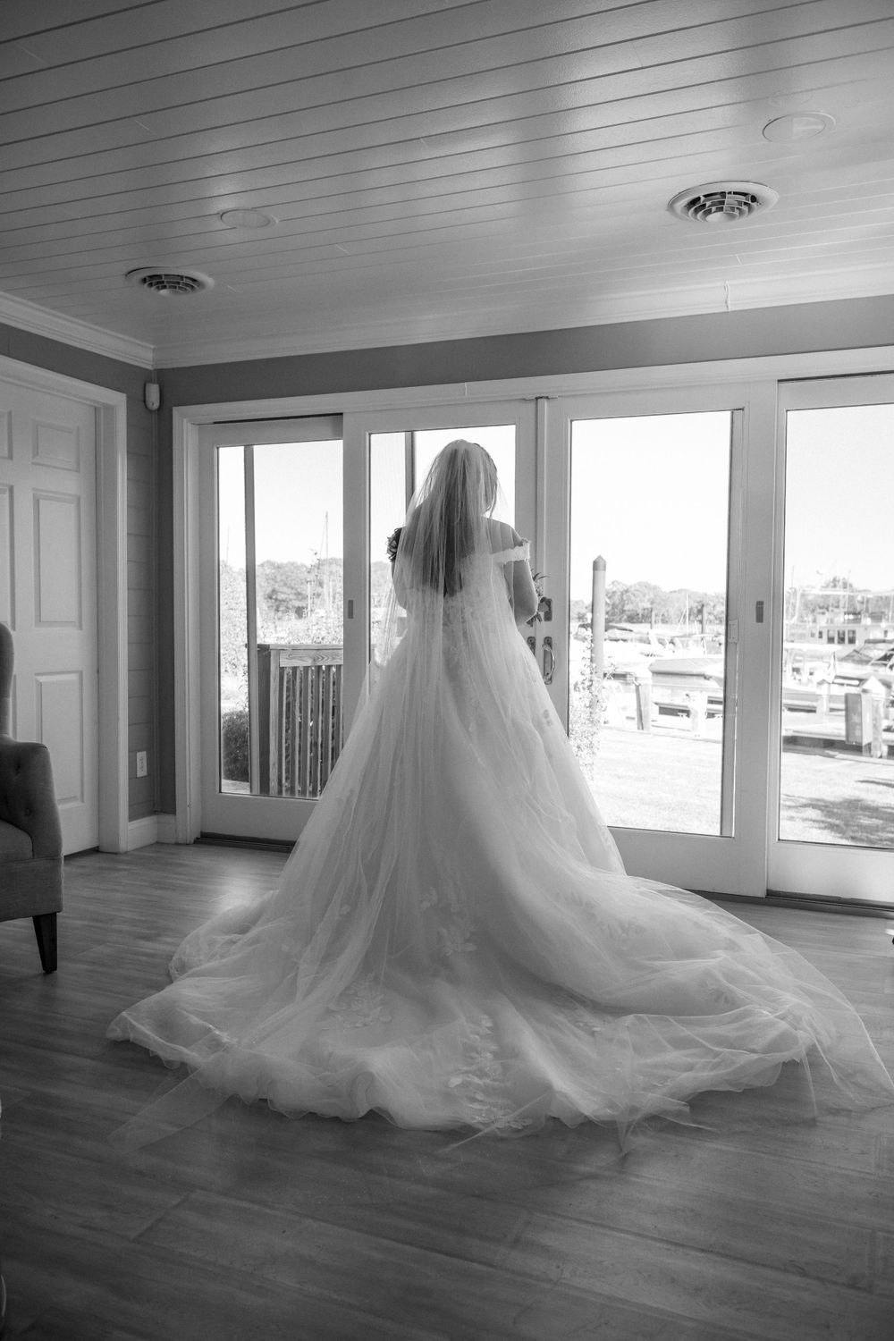 Wedding dress with flowing train silhouetted against bright windows overlooking marina.