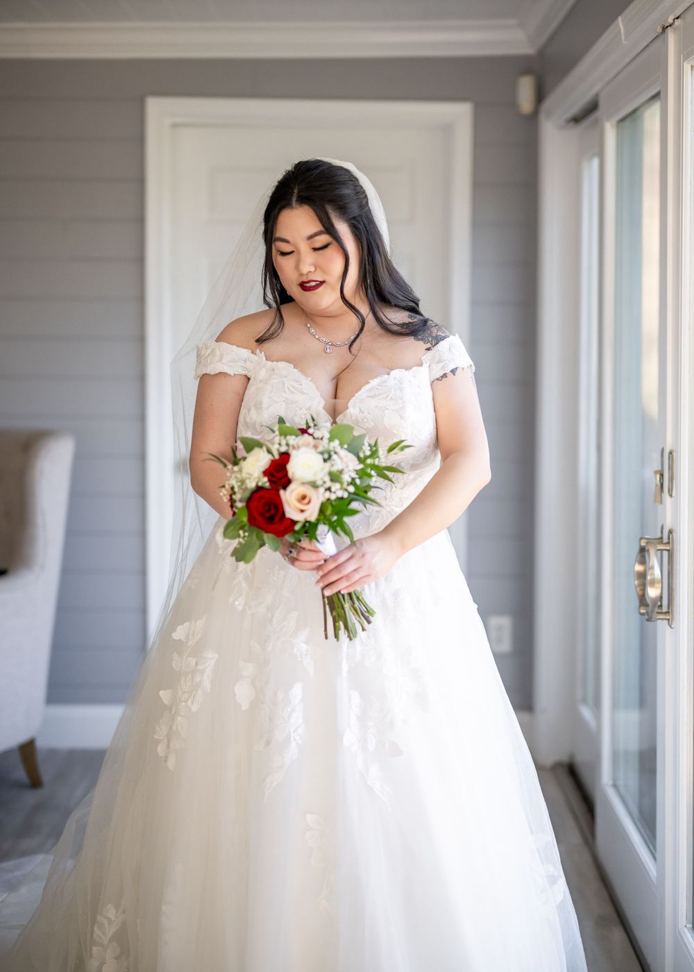 A bride wearing an off-shoulder white wedding gown holds a red and white bouquet in a bridal portrait.