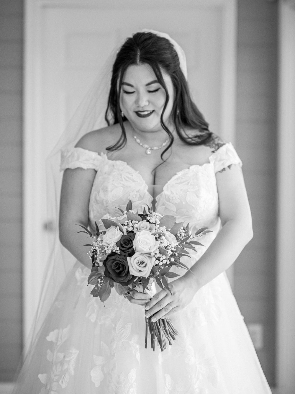 A black and white portrait of a bride in a lace off-shoulder gown holding her wedding bouquet.