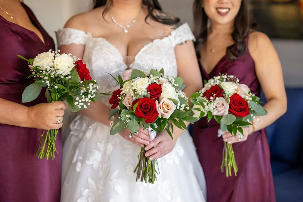 Three matching wedding bouquets with red roses and white flowers held against burgundy and white dresses.