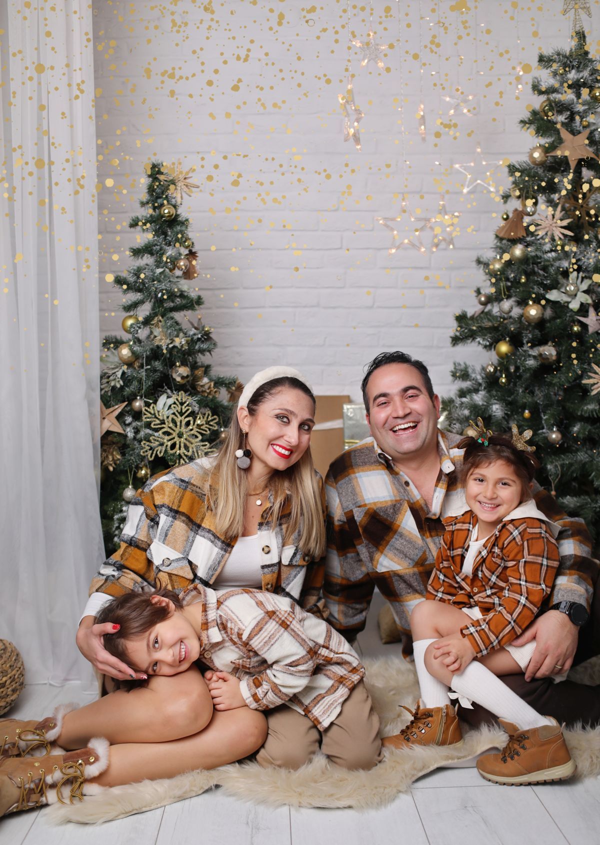 A family wearing matching plaid flannel shirts poses for a Christmas photo with decorated trees.
