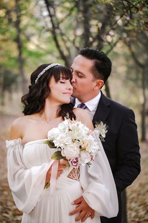 Romantic wedding portrait with couple embracing among trees while holding white floral bouquet.