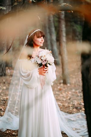 Bride in flowing white dress with bishop sleeves holds pink bouquet during autumn forest photoshoot.