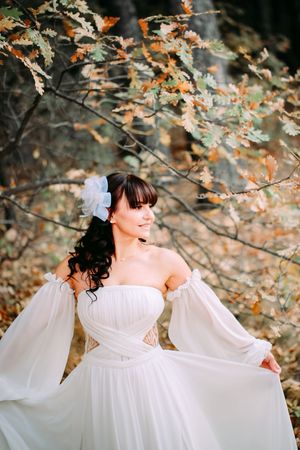 Bride in flowing white dress poses elegantly against autumn foliage.