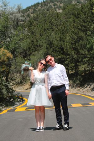 A couple in casual white outfits poses playfully on a mountain road lined with evergreen trees.