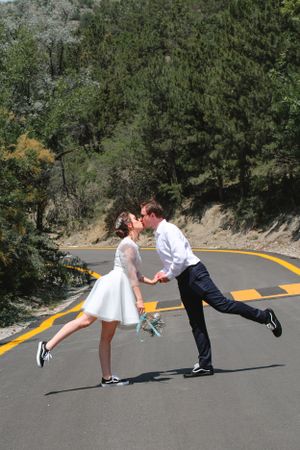 Playful wedding photo of couple kissing while balanced on one leg on a mountain road.