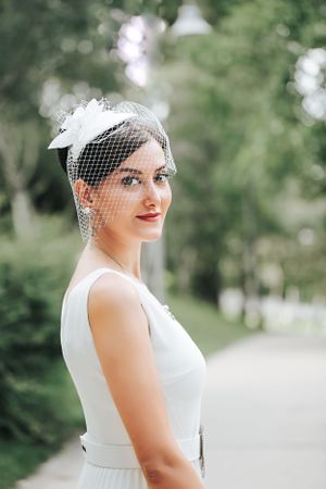 Elegant bride in a white dress wearing a vintage-style birdcage veil in an outdoor garden setting.