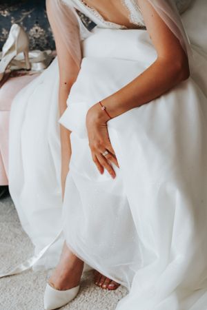 Close-up of a hand wearing delicate jewelry against a white wedding dress and high heels.