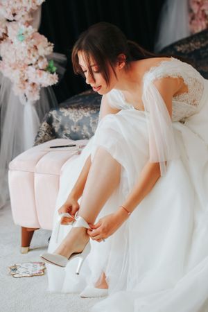 Bride in delicate lace dress puts on white wedding shoes while sitting on pink ottoman.