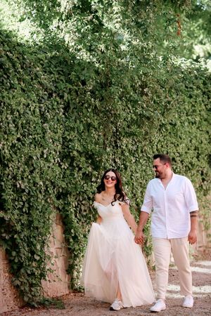 A wedding couple in white outfits walking together along an ivy-covered wall.