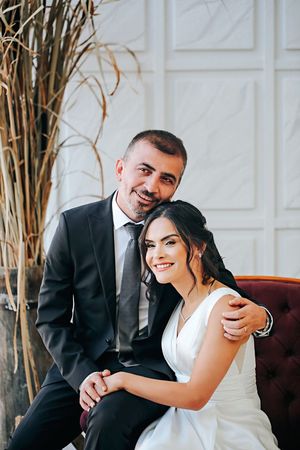 Elegant couple poses for wedding portraits in formal attire against a white interior backdrop.