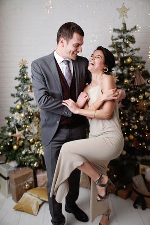 A couple laughs together in front of a decorated Christmas tree during holiday celebrations.