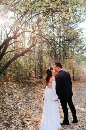 A wedding couple shares an intimate moment under sunlit trees on a forest path.