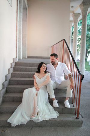 Couple in white sitting on stone steps with classic architecture in background.