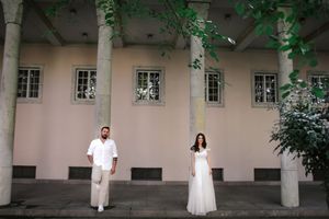 Wedding couple in white outfits standing apart against pink building with columns and windows.