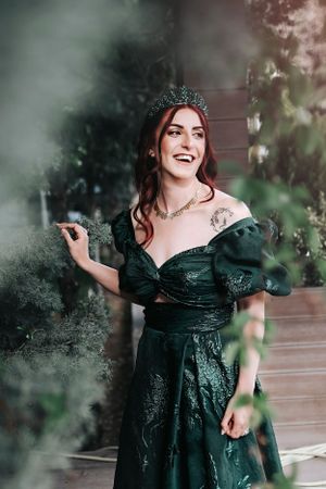A woman in an emerald green gown with puff sleeves and tiara poses elegantly in a dreamy outdoor setting.