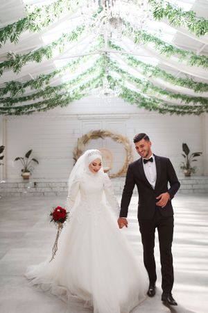 Wedding couple walks in white venue decorated with hanging greenery and crystal chandelier.