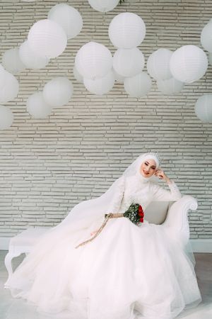 Bride in white gown poses on chaise lounge under paper lantern decor.