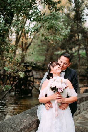 A romantic moment between newlyweds holding a pink bouquet by a stream.