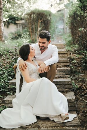 A romantic wedding couple sits together on rustic stone steps surrounded by lush garden greenery.