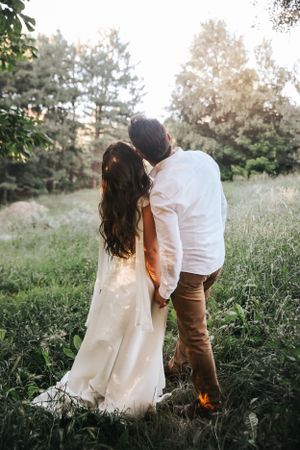 Couple in casual wedding attire standing together in sunlit meadow with forest backdrop.