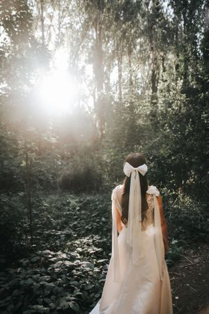 Back view of bride in white dress with long veil and bow walking through sunlit forest.