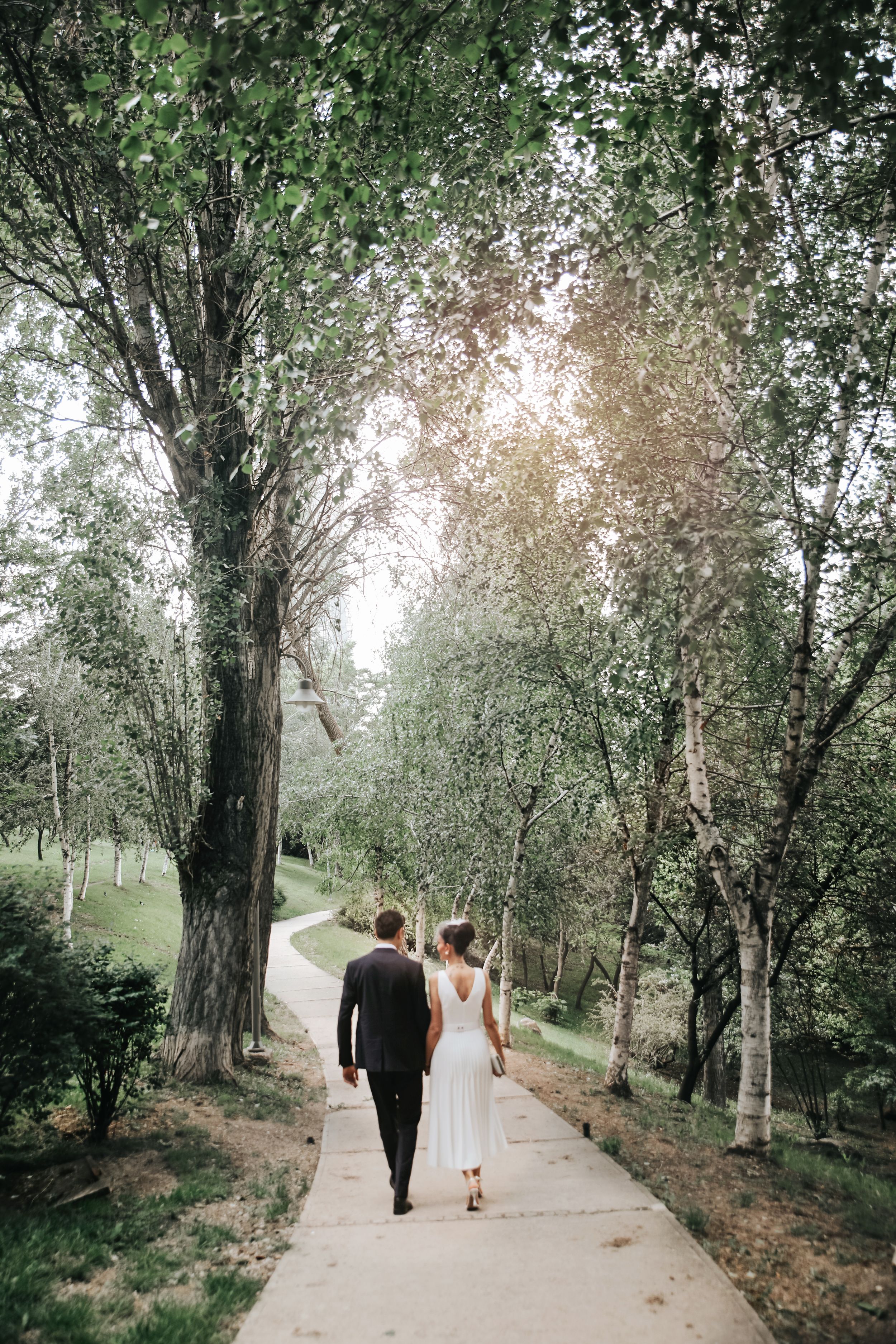 Newlyweds walking hand in hand down a tree-lined path with sunlight filtering through branches.