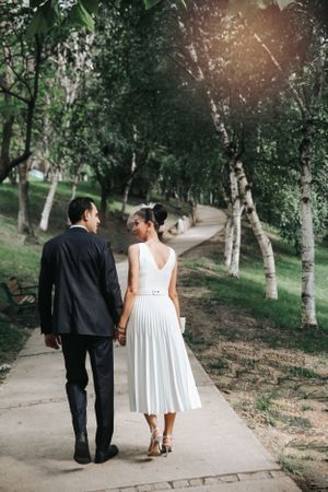Couple walking hand in hand along a tree-lined path in wedding attire.