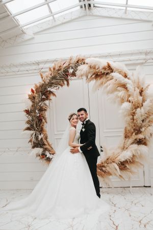 Newlyweds pose under a stunning arch decorated with dried pampas grass and botanical elements.