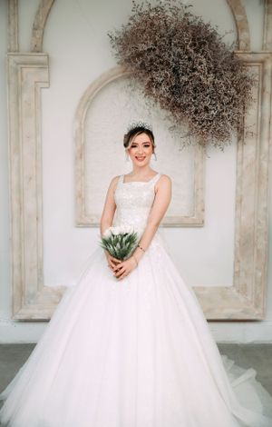 A bride in a classic white ball gown holds a small succulent bouquet in front of an ornate archway with dried florals.