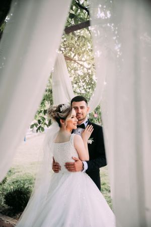 A romantic wedding portrait through flowing white curtains in a garden setting.