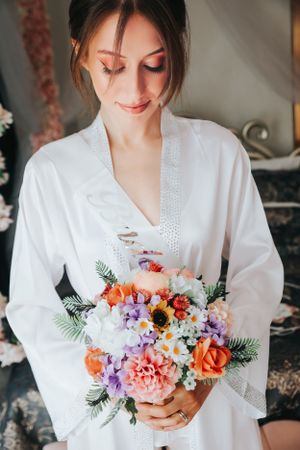 Bride in white robe holds vibrant bouquet of mixed flowers during wedding preparations.