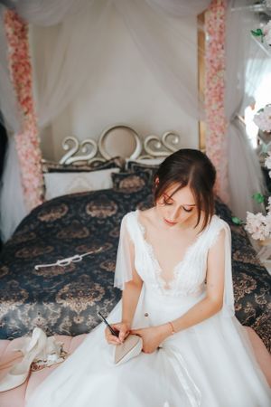 A bride in white dress sits on ornate bed preparing for wedding day surrounded by soft decor.