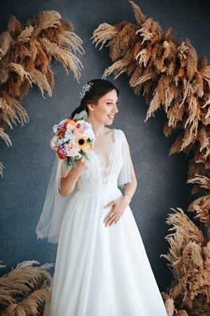 Elegant bridal portrait with dried pampas grass arch and colorful bouquet against dark backdrop.