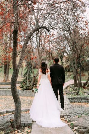 A couple in wedding attire walking along a cobblestone path surrounded by autumn trees.