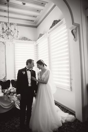 Elegant black and white wedding photo in a formal room with grand windows and chandelier.