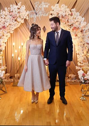 Elegant couple standing beneath ornate floral arch with golden backdrop at indoor venue.
