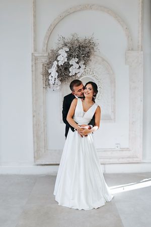 Couple shares intimate moment under ornate white archway with floral decorations.