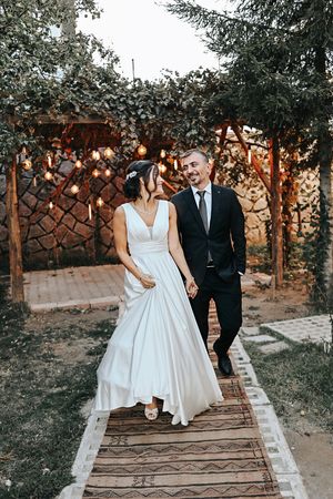 Couple walks along wooden pathway under string lights and garden trellis during wedding celebration.