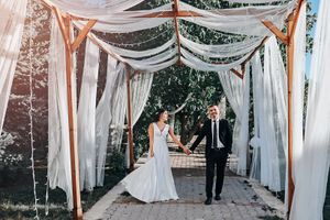 Wedding couple walks through elegant outdoor corridor draped with white fabric and string lights.